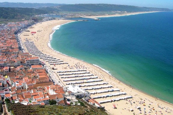 Aerial view of a crowded beach with umbrellas and adjacent town buildings along a curved coastline.