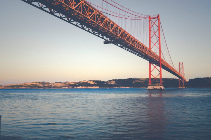 Red suspension bridge over calm water at sunset with land in the background.