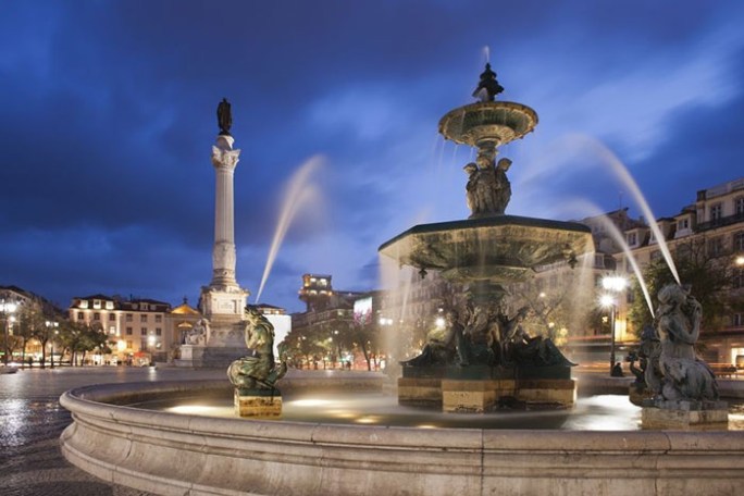 Lisbon square with illuminated fountain and statue at night, under a cloudy sky.