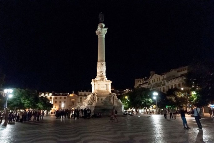 Night scene of a lit monument surrounded by people in a plaza with patterned pavement.