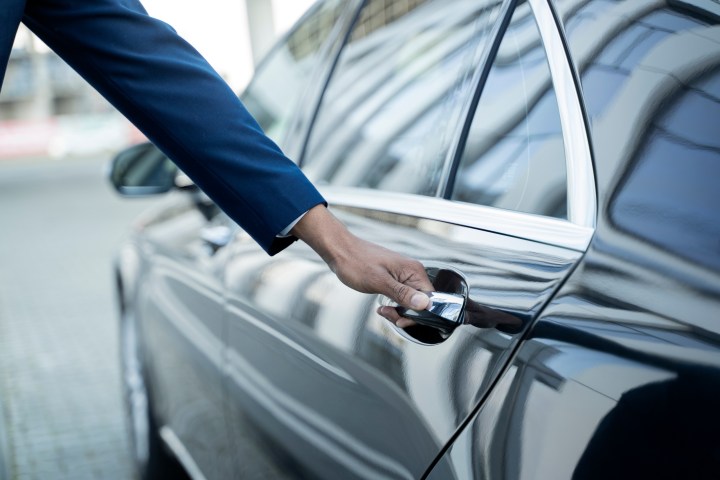 Person in a blue suit opening a black car door on a sunny day.