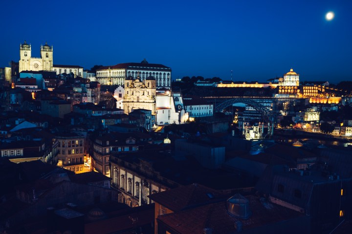 Night view of a city with illuminated historic buildings and a bridge under a bright moon.