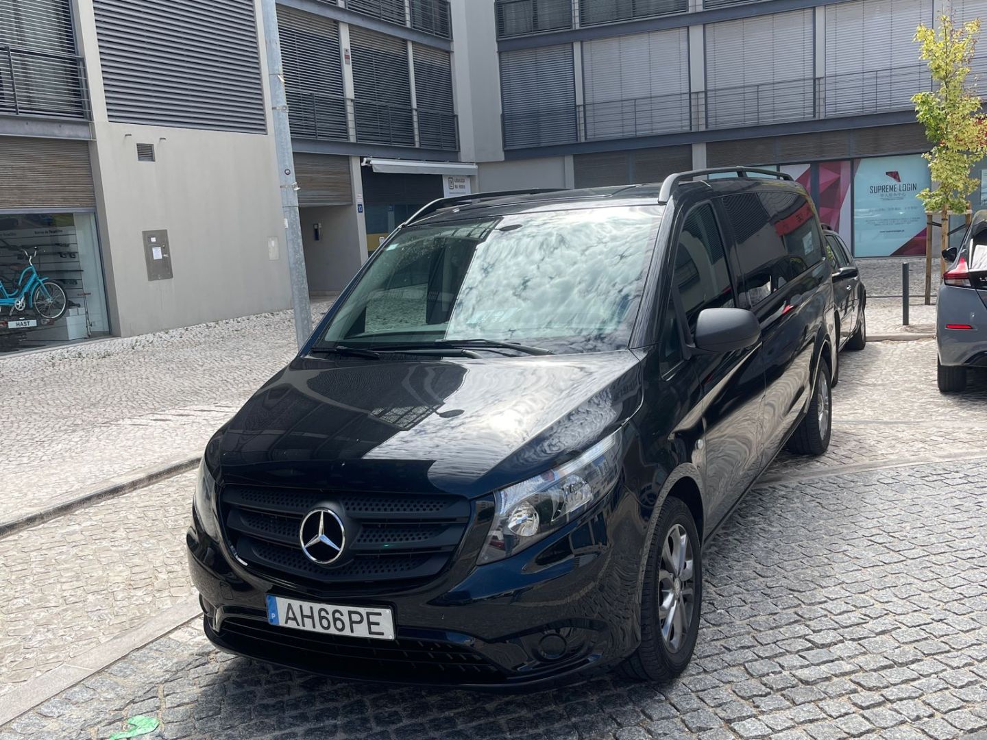 Black van parked on cobblestone street between modern buildings.