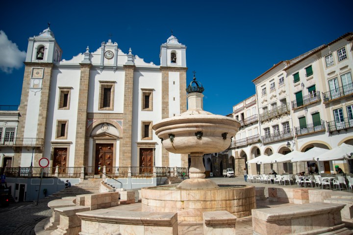 Historic square with a large fountain and a church under a clear blue sky.