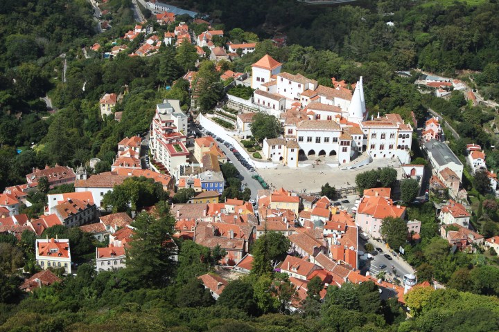 Aerial view of a town with red-roofed buildings and a white palace amidst lush greenery.