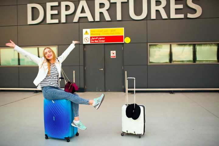 Woman sitting on suitcase, arms outstretched, in front of departures sign.