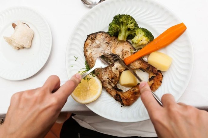 Person cutting grilled fish with vegetables and lemon on plate.