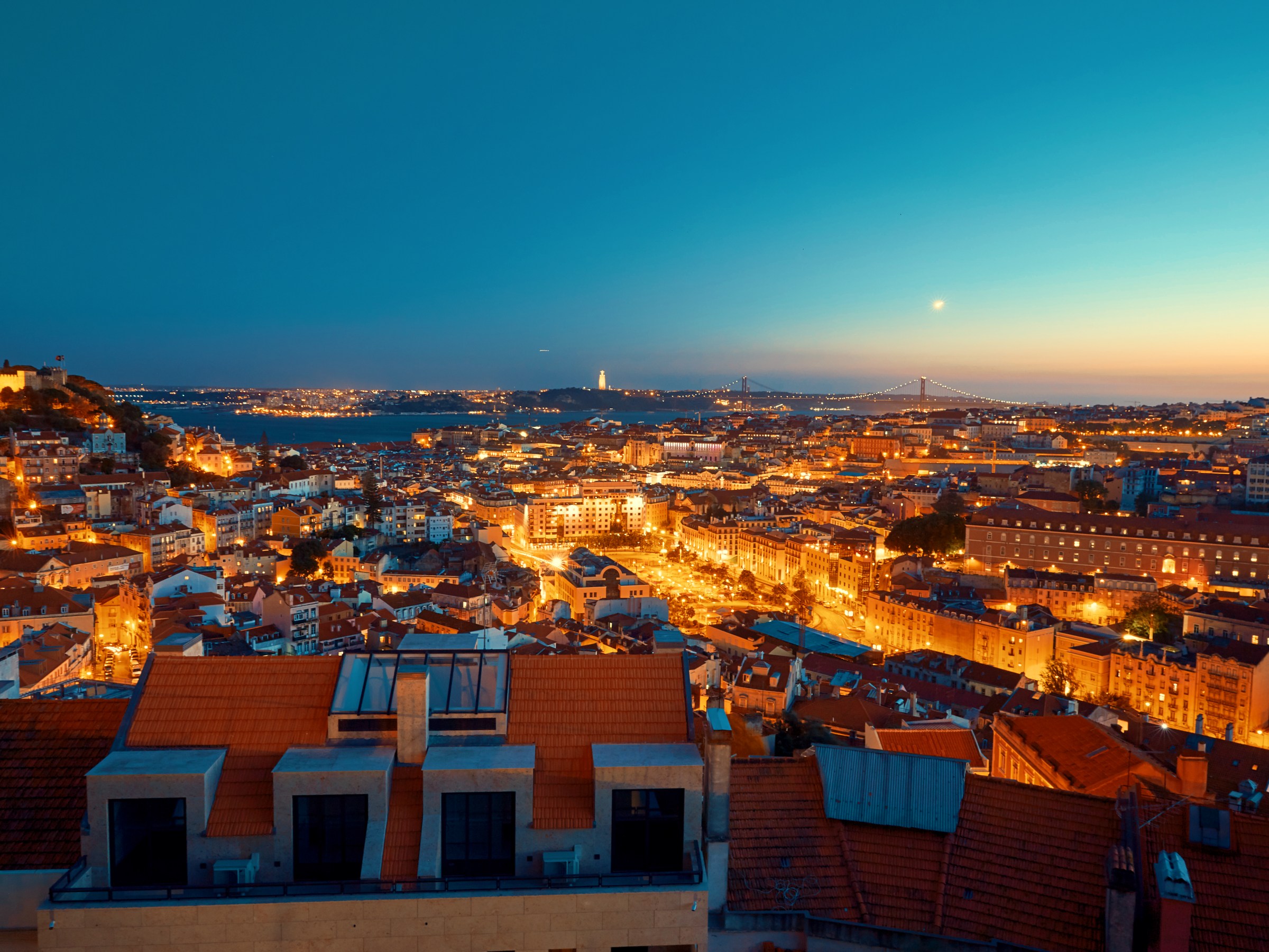 Night view of a cityscape with illuminated buildings and a bridge in the distance under a twilight sky.