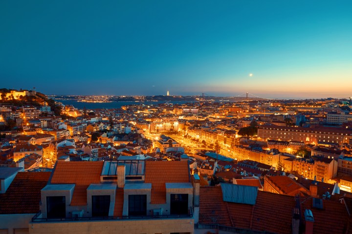 Night view of a cityscape with illuminated buildings and a bridge in the distance under a twilight sky.