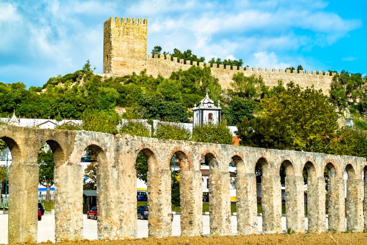 Stone aqueduct and medieval castle on a hill under a partly cloudy sky.