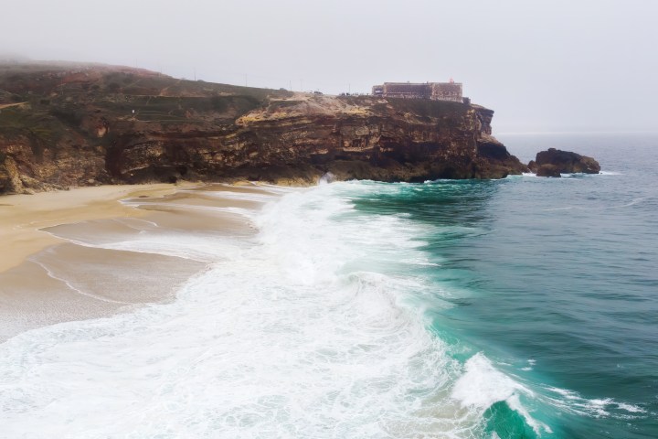 Coastal cliff with waves hitting the shore, topped by a building under a cloudy sky.