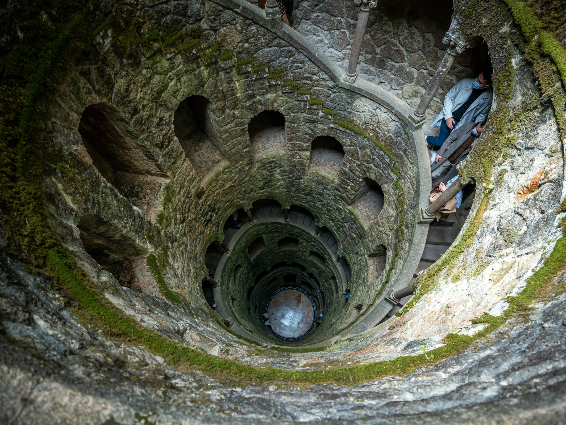 Looking down a spiral stone staircase with people, surrounded by mossy stone walls.