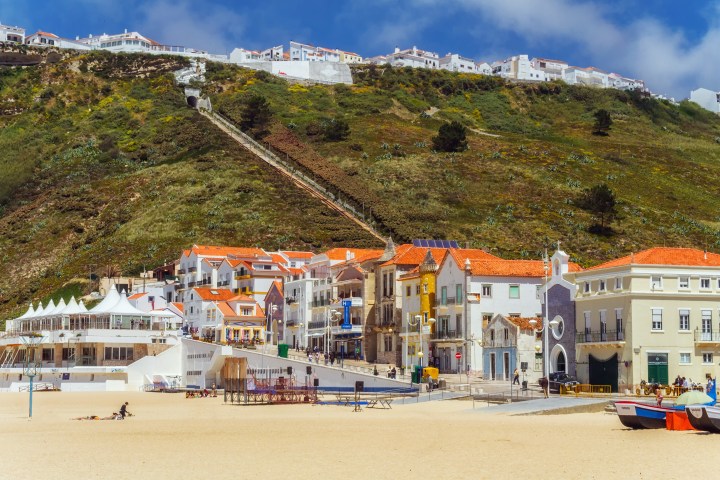 Beachfront village with colorful rooftops and hillside in the background under a blue sky.