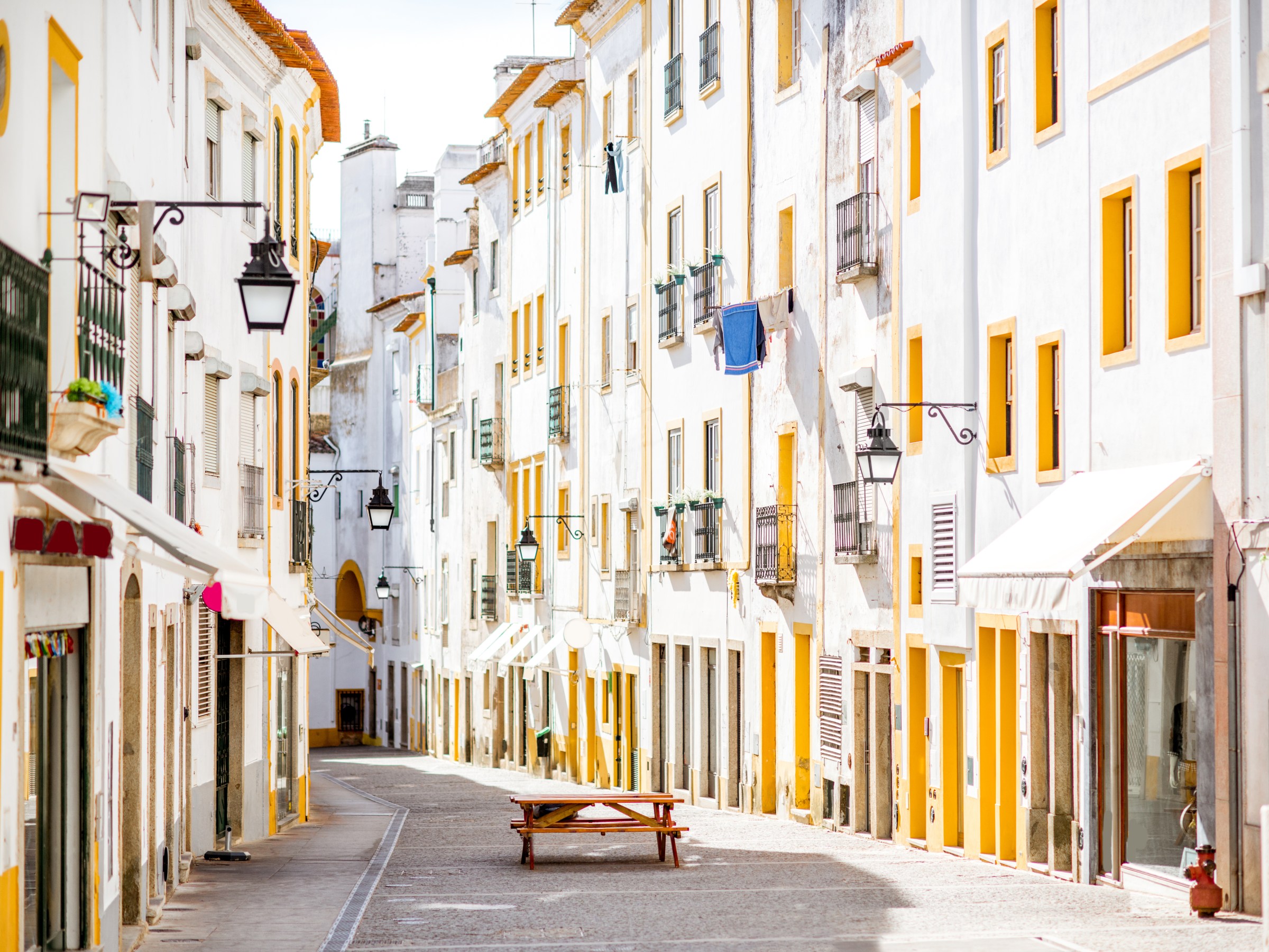 Narrow street with white buildings and orange accents; empty tables along the path under vintage lanterns.