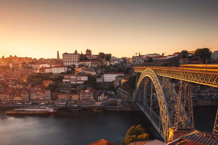 Cityscape with a bridge over a river at sunset, with buildings and rooftops in the foreground.