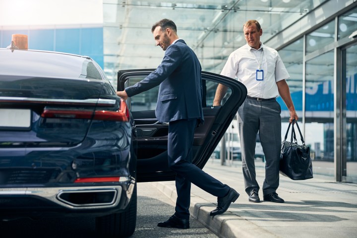 Man in suit entering black car while another man holds a bag nearby.